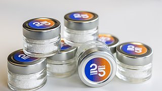 Acht kleine, runde Gläser mit silbernem Deckel, gefüllt mit weißem Salz. Auf jedem Glas ist ein Etikett mit der Aufschrift '25 Jahre dena' in Blau und Orange.