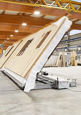Person richtet maschinell ein vorgefertigtes Hausfassadenelement in einer Werkhalle auf.