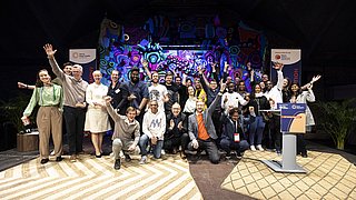 Gruppenfoto der Gewinner des SET Award 2023 auf Bühne mit buntem Hintergrund, einige Personen jubeln mit erhobenen Armen