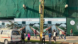 Menschen gehen an einer Straße mit einem weißen Kleinbus vorbei, im Hintergrund eine grüne Bauplane mit Werbetafeln und einem gelben Mast in der Bildmitte