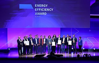 Gruppenszene von 14 Personen auf einer Bühne, die Urkunden für den Energy Efficiency Award 2022 halten. Im Hintergrund steht der Schriftzug 'ENERGY EFFICIENCY AWARD by dena'.