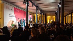 Drei Personen auf Bühne bei Start-up Energy Transition Festival, Publikum sitzt in großem Saal mit Säulen