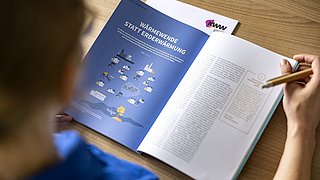 Person liest eine Broschüre mit dem Titel 'Wärmewende statt Erderwärmung' und hält dabei einen Stift in der Hand.