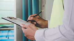 Nahaufnahme von einem Oberkörper und Händen, die ein Klemmbrett mit einem Formular halten. Mit einem Stift schreibt die Person ins Formular.