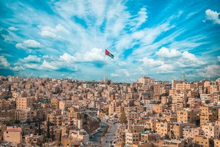 Panoramablick auf die Stadt Amman mit jordanischer Flagge, vor bewölktem Hintergrund.