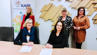 Fünf Personen in einem Büro bei der Unterzeichnung einer Kooperationsvereinbarung zwischen KAPE und dena, zwei sitzen am Tisch mit Dokumenten, drei stehen dahinter vor einer KAPE-Werbetafel.