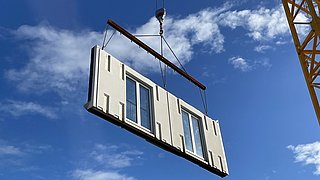 Betonfassadenelement mit Fenstern wird von einem Kranhaken vor blauem Himmel gehoben