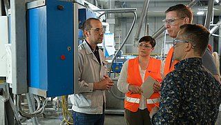 Eine Werksbesichtigung: Ein Mann in Arbeitskleidung erklärt einer Gruppe in Schutzbrillen und Warnwesten eine Maschine in einer Industriehalle.