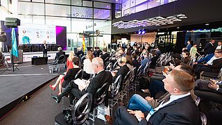 Die Konferenz "Tag der deutschen Wirtschaft in Kasachstan" findet in einem hellen großen Saal statt. Eine Rednerin steht auf der Bühne vor einer großen Präsentationsleinwand, während zahlreiche Teilnehmende in mehreren Reihen auf transparenten Stühlen sitzen und zuhören. Einige Personen tragen Headsets. Im Hintergrund sind Technikpersonal, Kameras und Beleuchtung zu sehen, die für die Veranstaltung eingesetzt werden.