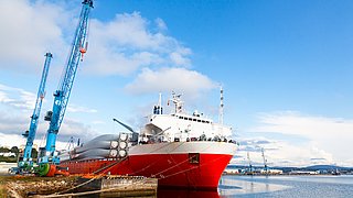 Am Pier eines Seehafens festgemachtes Transportschiff, beladen mit Windgeneratorflügeln, in der Nähe stehen Hafenkräne.