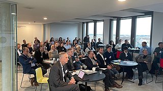 Mehrere Personen sitzen in einem modernen Konferenzraum an Tischen und hören einem Vortrag zu.