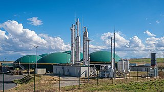Biogasanlage mit mehreren großen grünen Kuppeln und hohen silbernen Industrieanlagen unter blauem Himmel mit Wolken