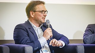 Zu sehen ist ein Mann mit Mikrofon in der Hand, der spricht und gestikuliert.