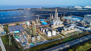 Luftaufnahme einer Industrieanlage mit mehreren silbernen Tanks, rauchenden Schornsteinen und einem großen Solarfeld im Hintergrund.