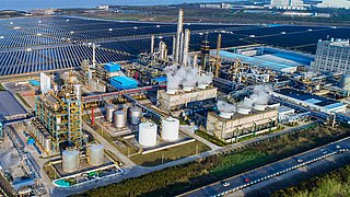 Luftaufnahme einer Industrieanlage mit mehreren silbernen Tanks, rauchenden Schornsteinen und einem großen Solarfeld im Hintergrund.