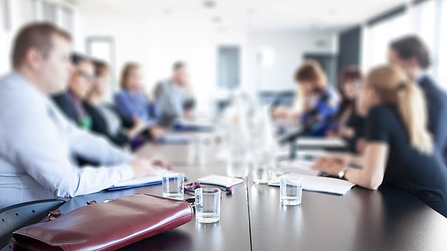 Mehrere Personen sitzen an einem langen Konferenztisch mit Gläsern und Unterlagen, unscharfer Hintergrund.