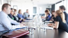 Mehrere Personen sitzen an einem langen Konferenztisch mit Gläsern und Unterlagen, unscharfer Hintergrund.