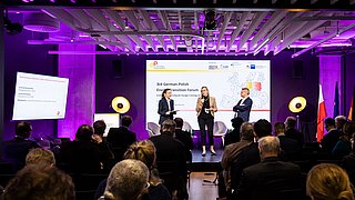Drei Personen auf einer Bühne vor Publikum in einem Konferenzraum mit lila Beleuchtung und Präsentationsbildschirm im Hintergrund