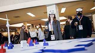 Frau mit Virtual-Reality-Brille steht vor einem großen, weißen Modell mit blauen und roten Elementen, im Hintergrund mehrere Personen in einem Konferenzraum.
