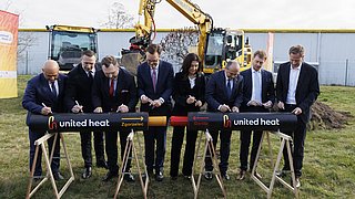 Sieben Personen in formeller Kleidung stehen hinter zwei schwarzen Rohren mit der Aufschrift 'united heat' und 'Zgorzelec' beziehungsweise 'Görlitz' und schreiben darauf, im Hintergrund ein Bagger auf einer Wiese