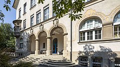 Eingang der Theodor-Heuss-Realschule mit drei Säulen und einem Treppenaufgang. Fenster mit weißen Rahmen und blauen Türen.