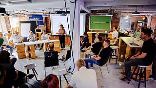 Ein moderner Coworking-Space mit Backsteinwänden: Eine Frau hält eine Präsentation vor einer Gruppe, die auf Stühlen und Hockern sitzt.
