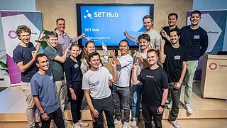 14 Personen stehen lächelnd auf einer Bühne und zeigen mit ihren Fingern auf einen Bildschirm hinter ihnen mit der Aufschrift SET Mentoring Kick-off