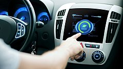 Ein Fahrer berührt den Touchscreen eines Autos, auf dem ein grünes 'ECO'-Symbol angezeigt wird. Das Armaturenbrett hat blaue Beleuchtung und verschiedene Bedienelemente.