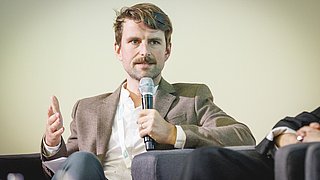 Zu sehen ist ein Mann der mit Mikrofon in der Hand spricht und gestikuliert.