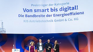 Drei Personen stehen auf einer Bühne vor einem Banner mit dem Text 'Preisträger der Kategorie Von smart bis digital! Die Bandbreite der Energieeffizienz KIS Antriebstechnik GmbH & Co. KG'. Eine Person hält eine Urkunde, eine weitere eine Trophäe.