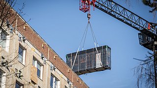 Ein von einem Kran gehaltenes vorgefertigtes Fassadenteil schwebt über dem Plattenbau.