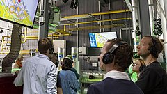  Personen sehen sich ein Video auf einem großen Bildschirm in einer Fabrik an.