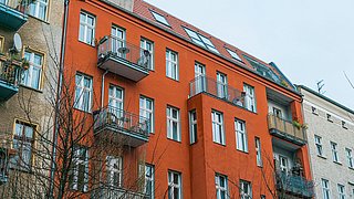Fassade eines orangefarbenen Mehrfamilienhauses mit Balkonen und Fenstern, umgeben von kahlen Bäumen.