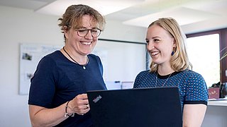 Zwei Frauen in einem Büro, die einen Laptop gemeinsam halten und sich unterhalten.