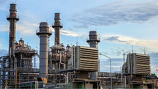 Es zeigt eine große Industrieanlage mit mehreren hohen metallischen Strukturen, darunter Schornsteine und zylindrische Türme, die wahrscheinlich Teil eines Kraftwerks oder einer Raffinerie sind. 