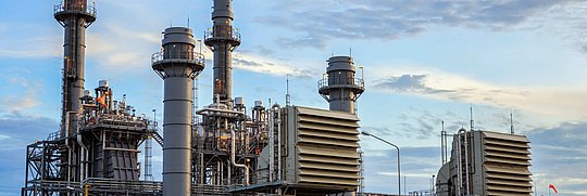 Es zeigt eine große Industrieanlage mit mehreren hohen metallischen Strukturen, darunter Schornsteine und zylindrische Türme, die wahrscheinlich Teil eines Kraftwerks oder einer Raffinerie sind. 