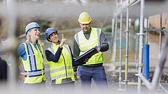 Drei Personen mit Schutzhelmen und Warnwesten stehen auf einer Baustelle, eine Person zeigt mit dem Finger nach oben, eine hält eine Bauzeichnung