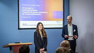Friederike Altgelt und Bastian Jakob stehen vor einer Präsentationsleinwand mit dem Titel 'H2-Hochlauf in Schlüsselindustrien: Potenziale für grünes Ammoniak in der Düngemittelindustrie', Publikum im Vordergrund.