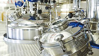 Zwei große, runde Edelstahltanks mit transparenten Deckeln und blauen Ventilen in einer industriellen Umgebung. Der Boden ist mit einem rautenförmigen Muster versehen.