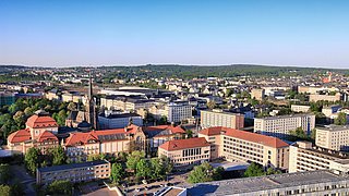 Luftaufnahme einer Stadt mit mehreren Gebäuden, darunter ein großes historisches Gebäude mit roten Dächern und ein hoher Kirchturm