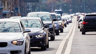Stau mit mehreren Autos auf einer Straße. Sichtbare Fahrzeuge sind in verschiedenen Farben, darunter Weiß, Schwarz und Silber.