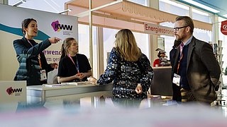 Vier Personen stehen an einem Messestand mit dem Schriftzug 'Energiesprong'. Eine Person zeigt mit ausgestrecktem Arm auf etwas außerhalb des Bildes.