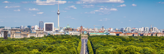 Panoramablick auf Berlin mit dem Tiergarten im Vordergrund, dem Brandenburger Tor in der Mitte und dem Fernsehturm sowie weiteren Gebäuden im Hintergrund unter blauem Himmel.