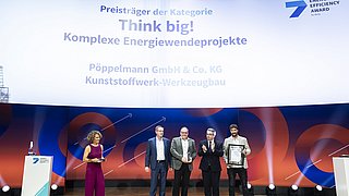 Fünf Personen auf Bühne bei Preisverleihung, eine Frau in rotem Jumpsuit, vier Männer in Anzügen, einer hält Urkunde. Im Hintergrund Text: 'Preisträger der Kategorie Think big! Komplexe Energiewendeprojekte Pöppelmann GmbH & Co. KG Kunststoffwerk-Werkzeugbau'.