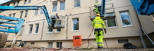 Montage eines vorgefertigten Fassadenelements an Mehrfamilienhaus, davor Kranfahrzeuge und Bauarbeiter
