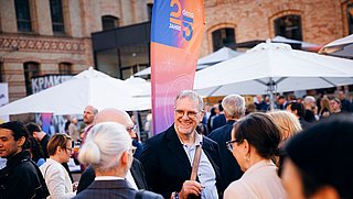 Das Bild zeigt Gäste bei der Jubiläumsfeier zum 25-jährigen Bestehen der dena. Im Hintergrund sind weiße Sonnenschirme und die Fassade der Königlichen Porzellanmanufaktur Berlin erkennbar.