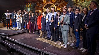 Gruppe von 15 Personen auf einer Bühne, einige tragen Anzüge, andere legere Kleidung. Im Hintergrund große dekorative Elemente und die Zahl '2025'.