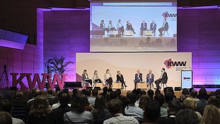 Podiumsdiskussion mit sieben Personen auf einer Bühne vor großem Publikum bei einer KWW-Konferenz in Halle in einem modern beleuchteten Saal.