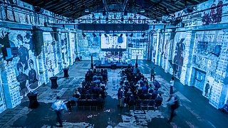 Die Eventlocation von innen, blau erleuchtet und mit dem Graphic recording an die Wand projiziert ist zu sehen.