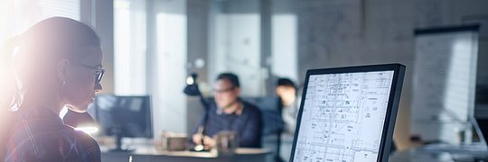 Person sitzt an einem Schreibtisch mit Tastatur und großem Monitor, auf dem technische Zeichnungen zu sehen sind, im Büro mit weiteren Personen im Hintergrund.